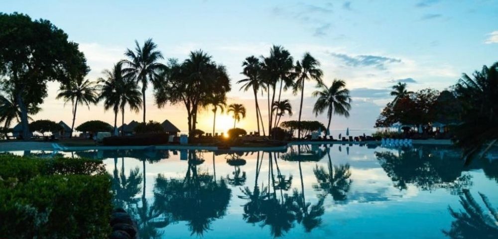 Tropical resort pool at sunset, with tall palm trees, thatched huts, and a glowing sky reflected in calm blue water; peaceful mood.
