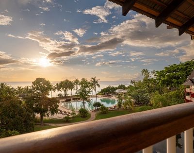 Golden sunset over a tropical resort, viewed from a balcony, with a pool, palm trees, lush gardens, and calm ocean beyond.