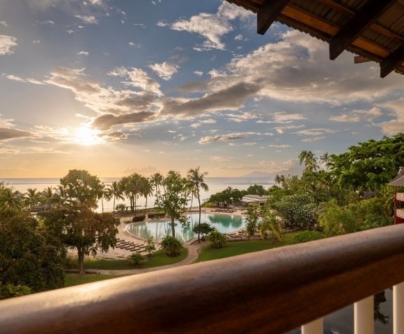Golden sunset over a tropical resort, viewed from a balcony, with a pool, palm trees, lush gardens, and calm ocean beyond.