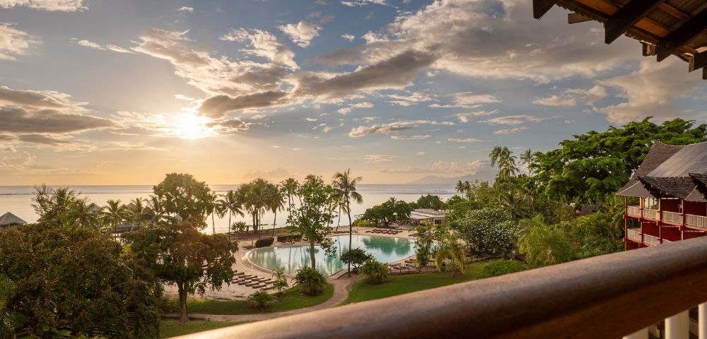 Golden sunset over a tropical resort, viewed from a balcony, with a pool, palm trees, lush gardens, and calm ocean beyond.