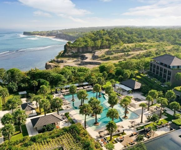 Aerial view of a tropical coastal resort with palm-lined pools, modern villas, lush greenery, and a rugged oceanfront cliff.