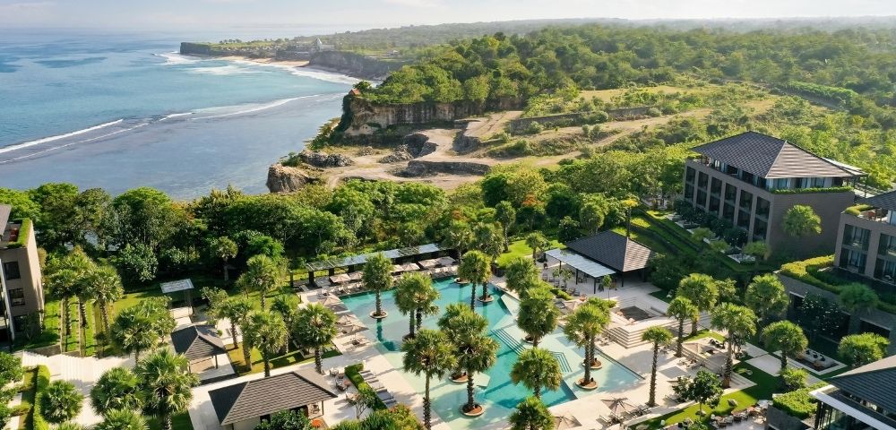 Aerial view of a tropical coastal resort with palm-lined pools, modern villas, lush greenery, and a rugged oceanfront cliff.