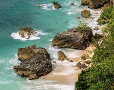 Turquoise waves wash over rugged limestone rocks and a small sandy cove, with swimmers offshore and tropical foliage along the coast.