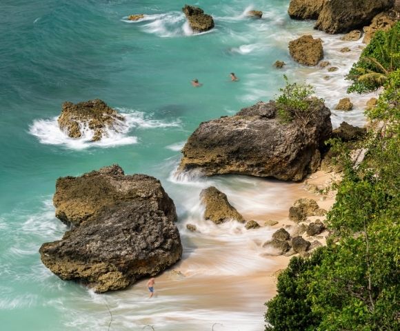 Turquoise waves wash over rugged limestone rocks and a small sandy cove, with swimmers offshore and tropical foliage along the coast.