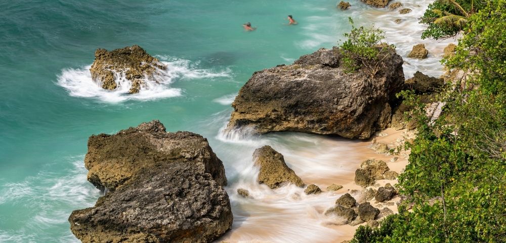 Turquoise waves wash over rugged limestone rocks and a small sandy cove, with swimmers offshore and tropical foliage along the coast.