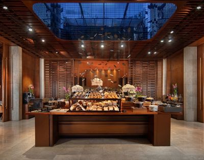Elegant restaurant buffet with breads, pastries, flowers, and warm pendant lights, framed by wood slats and a glass ceiling; inviting ambiance.