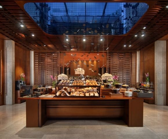Elegant restaurant buffet with breads, pastries, flowers, and warm pendant lights, framed by wood slats and a glass ceiling; inviting ambiance.