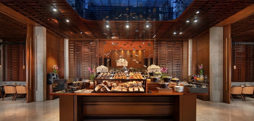 Elegant restaurant buffet with breads, pastries, flowers, and warm pendant lights, framed by wood slats and a glass ceiling; inviting ambiance.