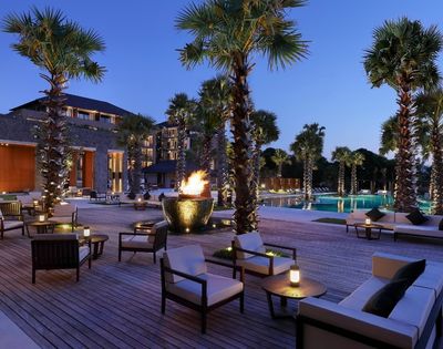 Luxury resort courtyard at dusk with palm trees, pool, glowing fire bowl, lounge chairs, and modern buildings creating a serene ambiance.
