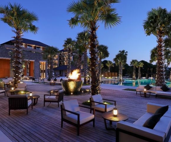 Luxury resort courtyard at dusk with palm trees, pool, glowing fire bowl, lounge chairs, and modern buildings creating a serene ambiance.