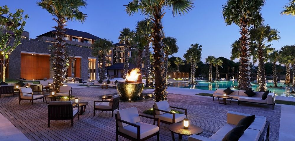 Luxury resort courtyard at dusk with palm trees, pool, glowing fire bowl, lounge chairs, and modern buildings creating a serene ambiance.
