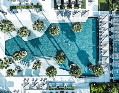Aerial view of a turquoise resort pool with geometric edges, palm islands, white loungers, and long shadows, creating a serene luxury atmosphere.