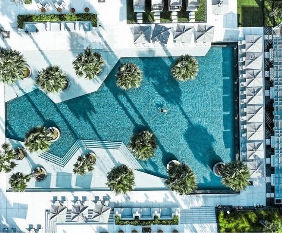 Aerial view of a turquoise resort pool with geometric edges, palm islands, white loungers, and long shadows, creating a serene luxury atmosphere.