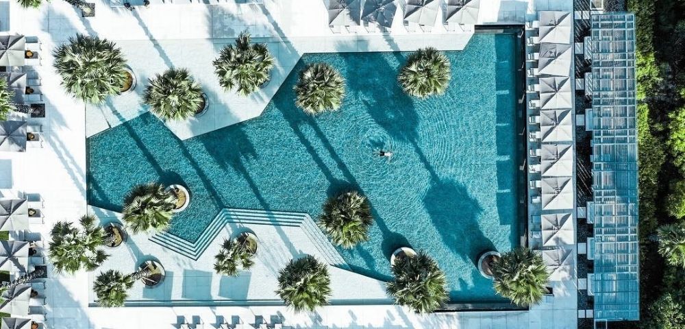 Aerial view of a turquoise resort pool with geometric edges, palm islands, white loungers, and long shadows, creating a serene luxury atmosphere.