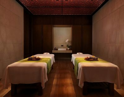 Dimly lit luxury spa room with two neatly made treatment tables, folded towels and trays, warm wood walls, and a calm, intimate atmosphere.