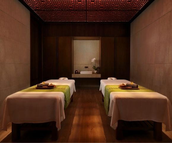 Dimly lit luxury spa room with two neatly made treatment tables, folded towels and trays, warm wood walls, and a calm, intimate atmosphere.
