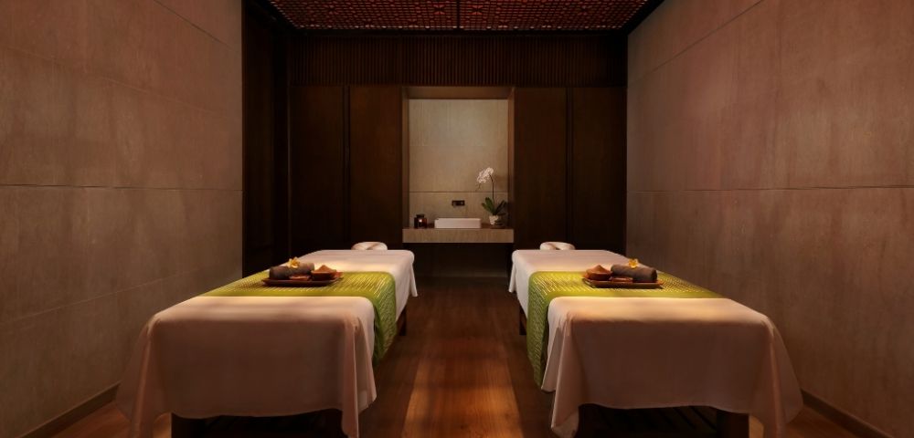Dimly lit luxury spa room with two neatly made treatment tables, folded towels and trays, warm wood walls, and a calm, intimate atmosphere.