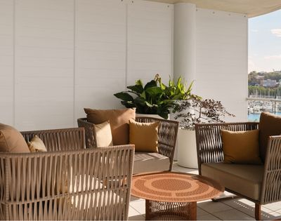 An outdoor seating area with woven wicker chairs and sofas adorned with brown and ochre cushions, arranged around a circular metal coffee table, with lush green plants in white planters, and a backdrop of a marina filled with boats and city buildings under a clear sky.