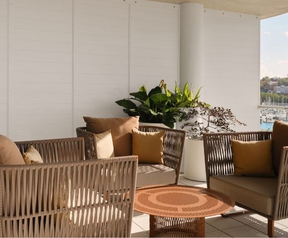 An outdoor seating area with woven wicker chairs and sofas adorned with brown and ochre cushions, arranged around a circular metal coffee table, with lush green plants in white planters, and a backdrop of a marina filled with boats and city buildings under a clear sky.