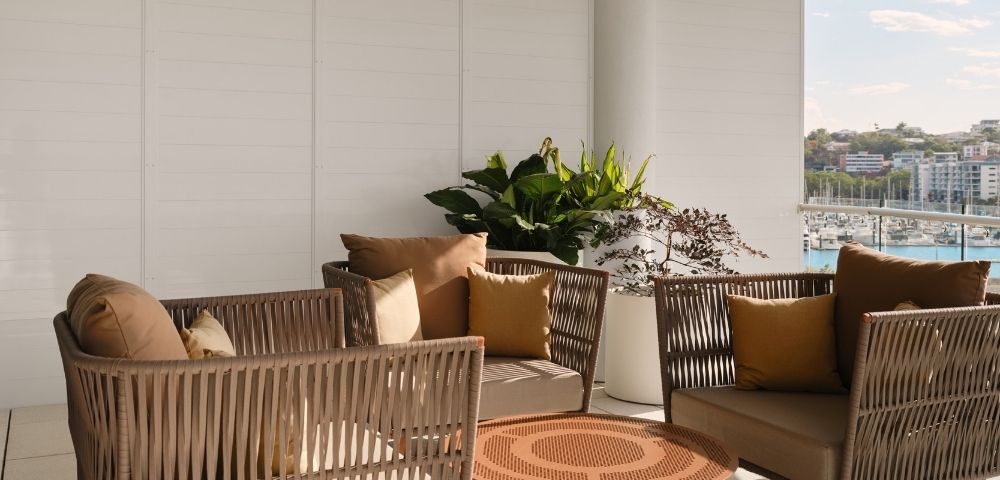 An outdoor seating area with woven wicker chairs and sofas adorned with brown and ochre cushions, arranged around a circular metal coffee table, with lush green plants in white planters, and a backdrop of a marina filled with boats and city buildings under a clear sky.
