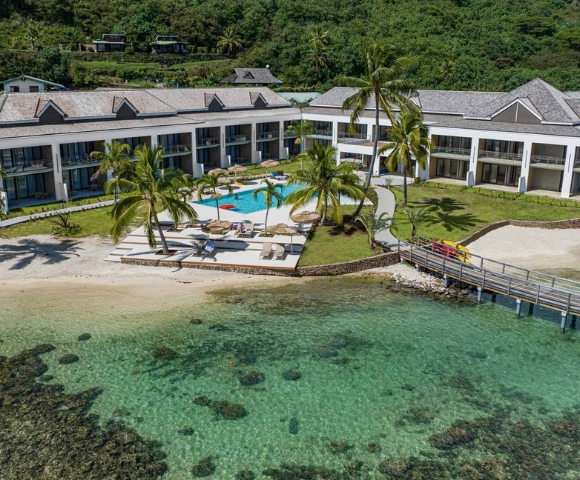 A modern waterfront resort overlooks clear turquoise water, with a long gray-roofed building, palm trees, a small dock, and a calm tropical atmosphere.