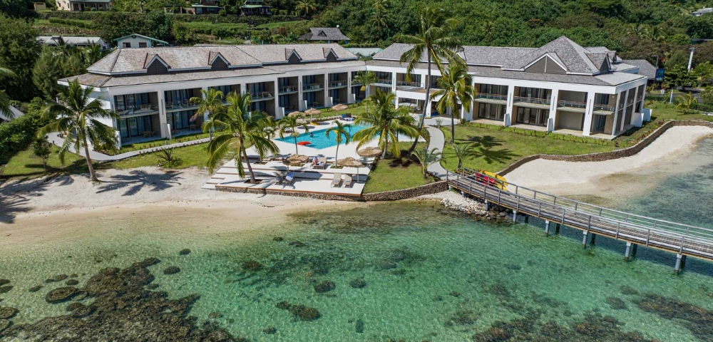A modern waterfront resort overlooks clear turquoise water, with a long gray-roofed building, palm trees, a small dock, and a calm tropical atmosphere.