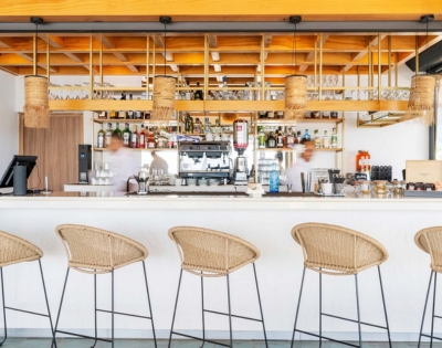 A warm modern café bar with woven tan pendant lamps and matching woven-seat stools, wood counters, and stocked shelves, conveying a calm, welcoming atmosphere.