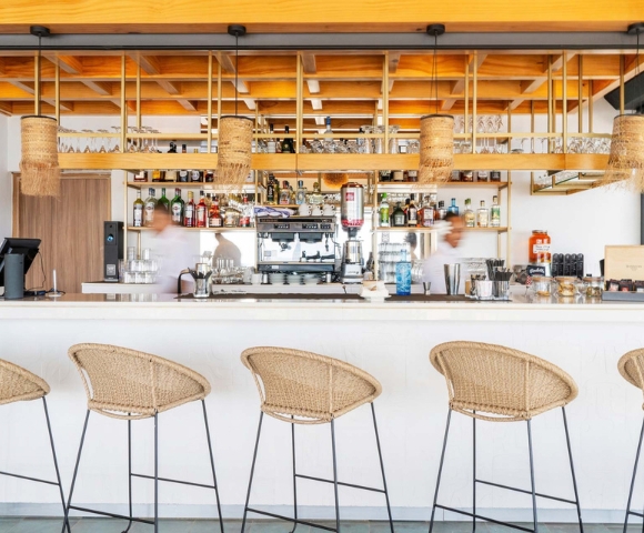A warm modern café bar with woven tan pendant lamps and matching woven-seat stools, wood counters, and stocked shelves, conveying a calm, welcoming atmosphere.