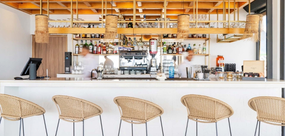 A warm modern café bar with woven tan pendant lamps and matching woven-seat stools, wood counters, and stocked shelves, conveying a calm, welcoming atmosphere.
