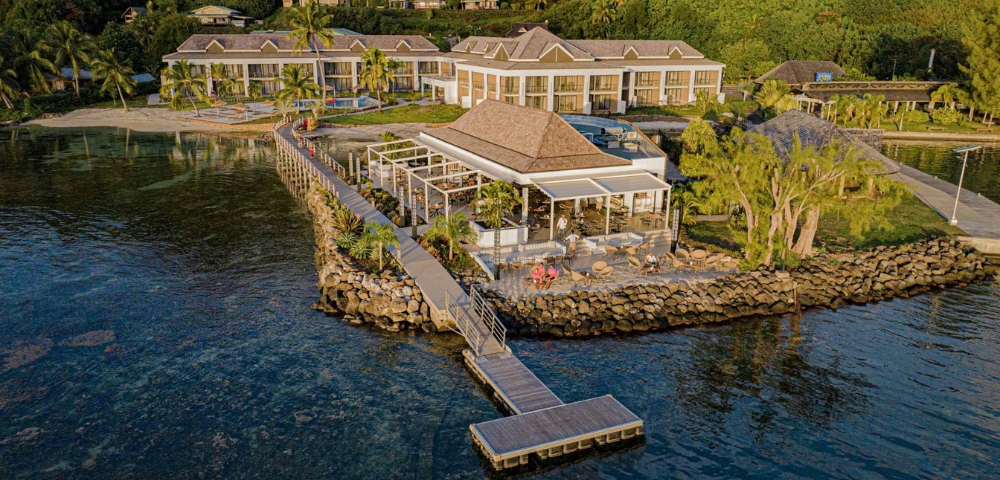 Architectural rendering of a modern waterfront home with stone retaining walls, patio, trees, and a long dock over calm blue water; serene setting.