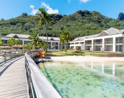 An architectural rendering of modern waterfront homes with palm trees, a wooden boardwalk, and a turquoise lagoon, set against green hills on a sunny day.