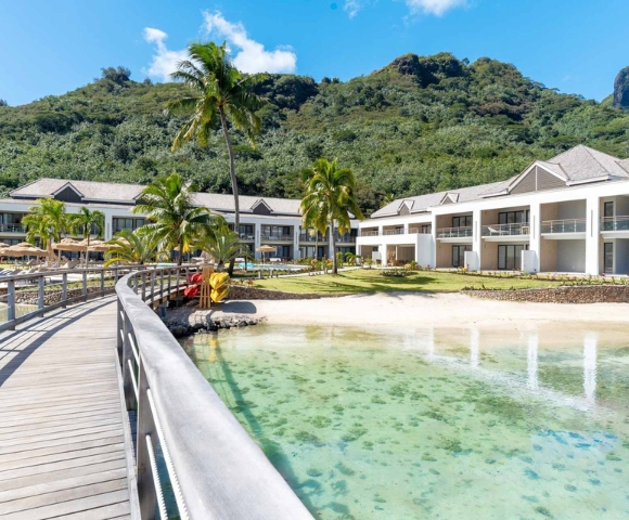 An architectural rendering of modern waterfront homes with palm trees, a wooden boardwalk, and a turquoise lagoon, set against green hills on a sunny day.
