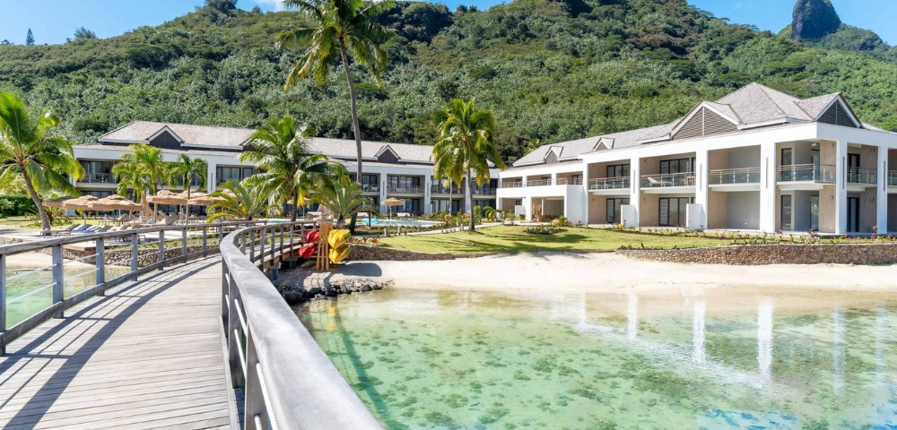 An architectural rendering of modern waterfront homes with palm trees, a wooden boardwalk, and a turquoise lagoon, set against green hills on a sunny day.