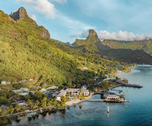 A tranquil tropical bay with a small waterfront village, palm-lined shore, and green mountain ridges beneath a clear blue sky; calm, scenic mood.