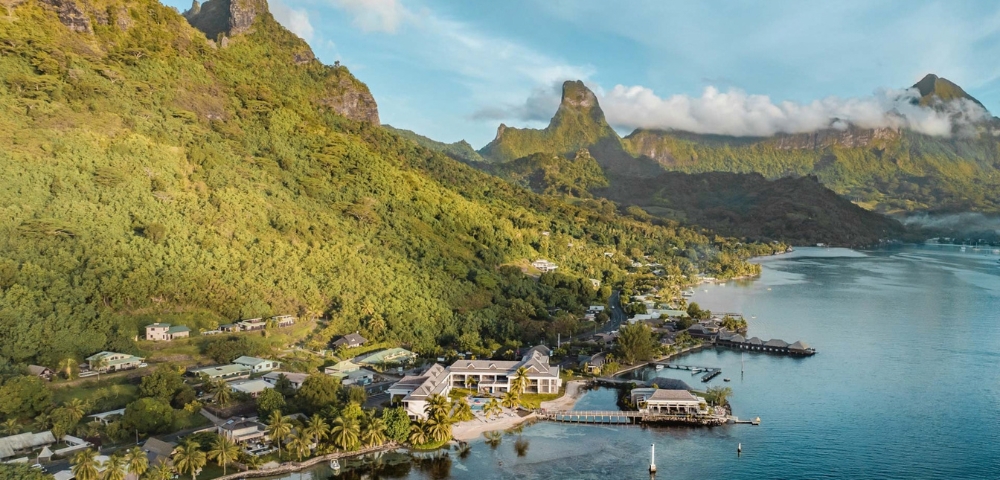 A tranquil tropical bay with a small waterfront village, palm-lined shore, and green mountain ridges beneath a clear blue sky; calm, scenic mood.