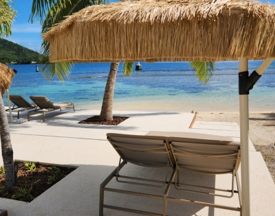 Two lounge chairs sit on a concrete patio facing a tropical beach with clear turquoise water, white sand, and palm trees, under a thatched umbrella.