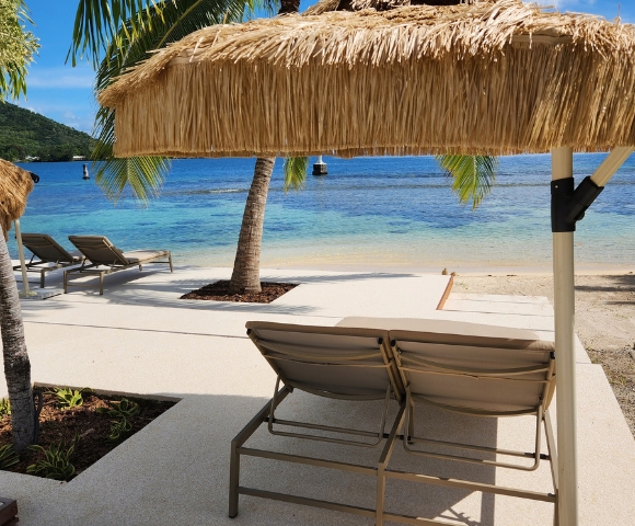 Two lounge chairs sit on a concrete patio facing a tropical beach with clear turquoise water, white sand, and palm trees, under a thatched umbrella.