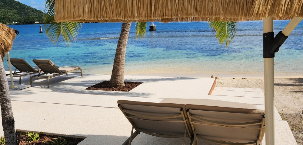 Two lounge chairs sit on a concrete patio facing a tropical beach with clear turquoise water, white sand, and palm trees, under a thatched umbrella.
