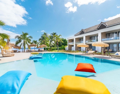 A bright resort pool with colorful floating loungers, palm trees, shaded cabanas, and ocean views under a blue sky, creating a calm vacation mood.
