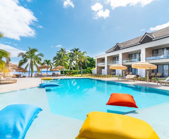 A bright resort pool with colorful floating loungers, palm trees, shaded cabanas, and ocean views under a blue sky, creating a calm vacation mood.