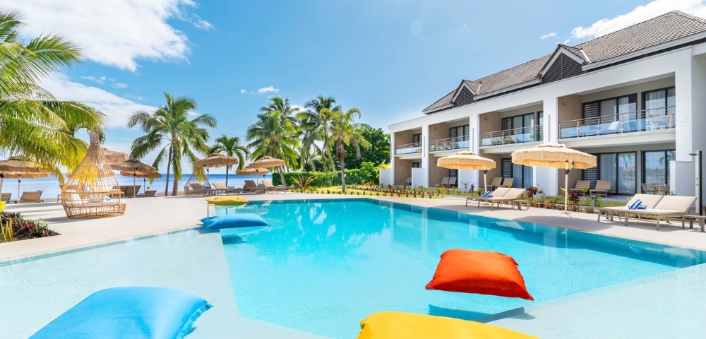A bright resort pool with colorful floating loungers, palm trees, shaded cabanas, and ocean views under a blue sky, creating a calm vacation mood.