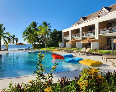 A resort swimming pool with inflatable loungers in red, blue, and yellow, adjacent to a modern building with balconies and palm trees, and a view of the ocean.