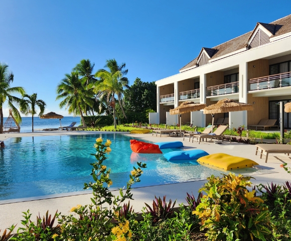 A resort swimming pool with inflatable loungers in red, blue, and yellow, adjacent to a modern building with balconies and palm trees, and a view of the ocean.