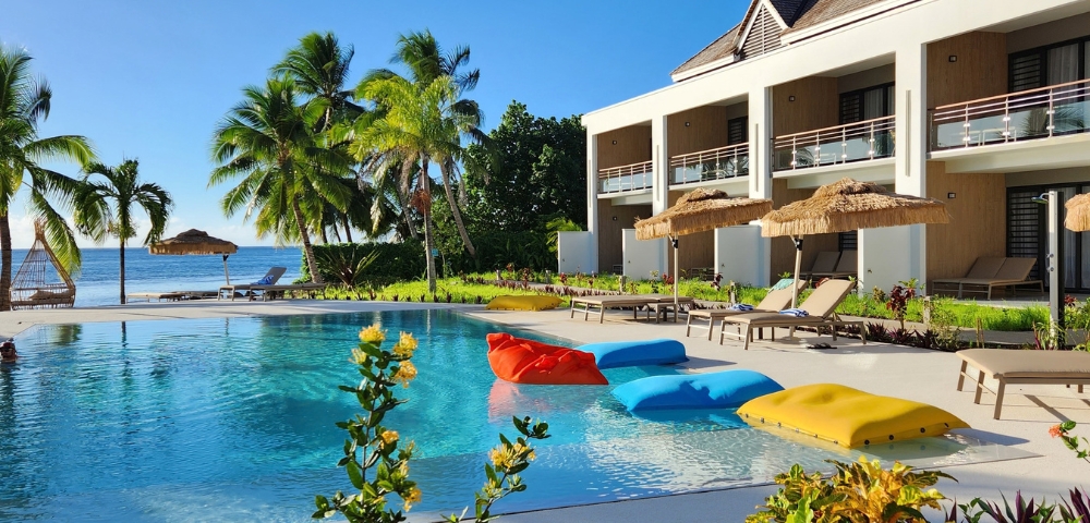 A resort swimming pool with inflatable loungers in red, blue, and yellow, adjacent to a modern building with balconies and palm trees, and a view of the ocean.