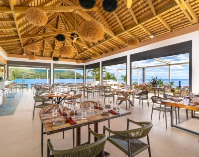 A bright modern restaurant dining room with neatly set tables, chairs, floor-to-ceiling windows overlooking greenery and water, and a dramatic wooden ceiling with woven pendant lights.