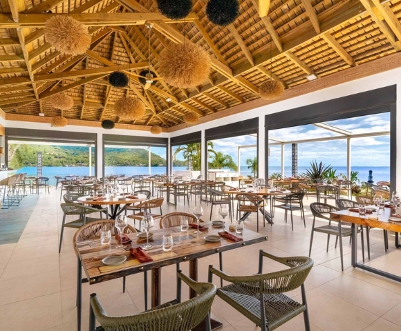 A bright modern restaurant dining room with neatly set tables, chairs, floor-to-ceiling windows overlooking greenery and water, and a dramatic wooden ceiling with woven pendant lights.