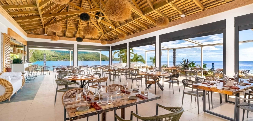 A bright modern restaurant dining room with neatly set tables, chairs, floor-to-ceiling windows overlooking greenery and water, and a dramatic wooden ceiling with woven pendant lights.