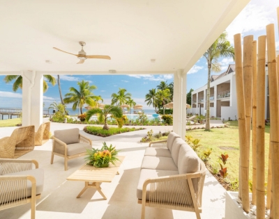 A covered patio with modern light-colored outdoor furniture faces a tropical resort scene with palm trees, a pool, and the ocean under a clear blue sky.