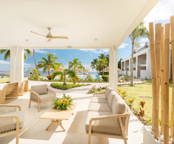 A covered patio with modern light-colored outdoor furniture faces a tropical resort scene with palm trees, a pool, and the ocean under a clear blue sky.