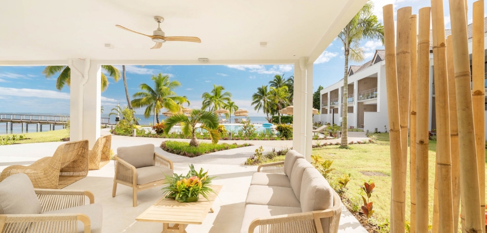 A covered patio with modern light-colored outdoor furniture faces a tropical resort scene with palm trees, a pool, and the ocean under a clear blue sky.